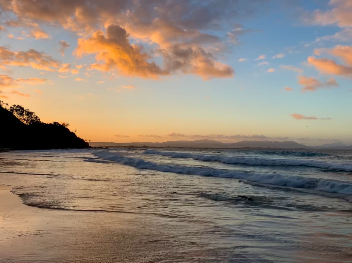 A serene beach scene with the sun setting or rising over the ocean, casting a warm glow on the waves and the sandy shore. The sky is filled with orange, pink, and blue hues with scattered clouds, and a silhouette of hills on the left side.